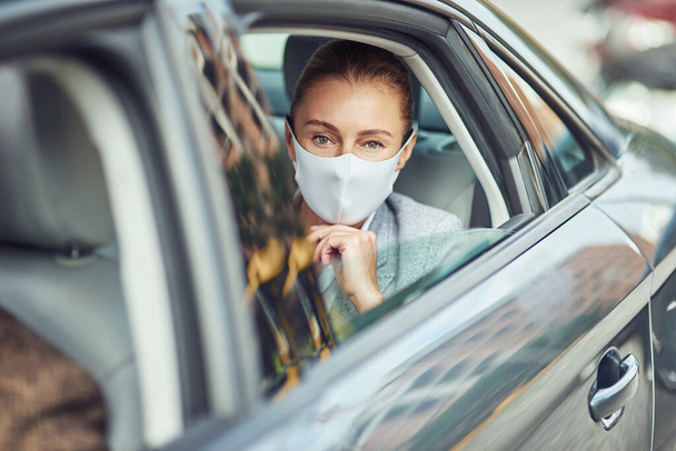 Кавказская деловая женщина в медицинской защитной маске сидит на заднем сидении в машине и смотрит в окно - Фото, изображение