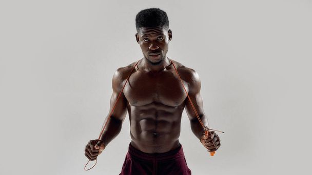 Young muscular african american man looking at camera, while holding jumping rope isolated over grey background. Sports, workout, bodybuilding concept - Foto, afbeelding