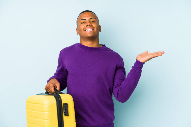 Young traveler man holding a suitcase isolated showing a copy space on a palm and holding another hand on waist. - Photo, Image