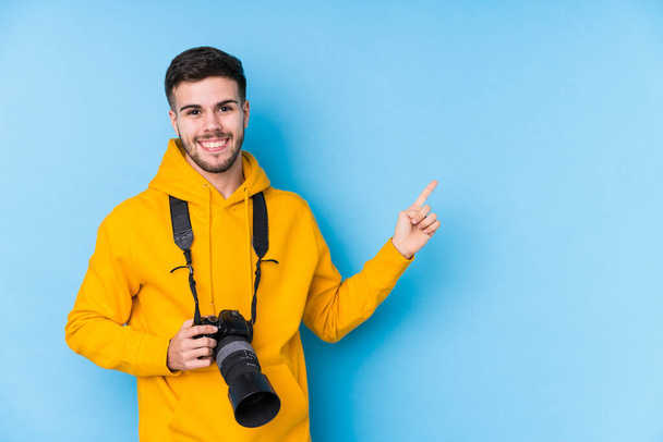 Jonge Kaukasische fotograaf man geïsoleerd glimlachend en wijzend opzij, iets laten zien op lege ruimte. - Foto, afbeelding