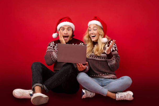 Young couple excited over Christmas discounts sitting with laptop on red background - Photo, Image