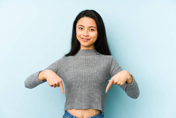Fiatal kínai nő elszigetelt kék háttér mutat lefelé ujjaival, pozitív érzés. - Fotó, kép