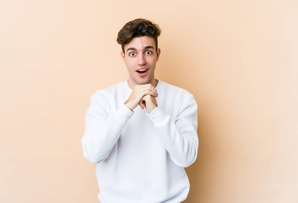 Young caucasian man isolated on beige background praying for luck, amazed and opening mouth looking to front. - Photo, Image