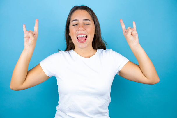 Νεαρή όμορφη γυναίκα πάνω από απομονωμένο μπλε φόντο φωνάζοντας με τρελή έκφραση κάνει ροκ σύμβολο με τα χέρια ψηλά - Φωτογραφία, εικόνα