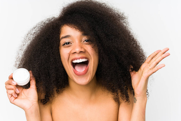 Junge Afro-Frau hält isoliert eine Feuchtigkeitscreme und feiert einen Sieg oder Erfolg - Foto, Bild