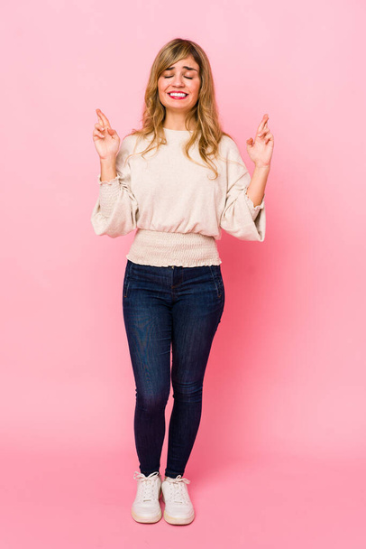 Young blonde caucasian woman standing over a pink background crossing fingers for having luck - Fotoğraf, Görsel