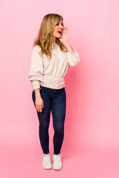 Young blonde caucasian woman standing over a pink background shouting and holding palm near opened mouth. - Foto, imagen