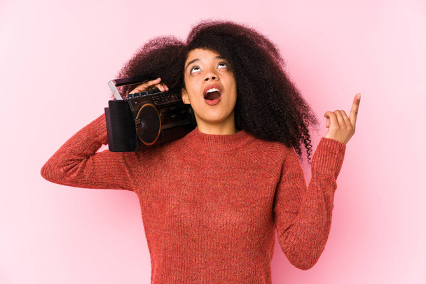 Joven mujer afro sosteniendo un cassete aislado apuntando hacia arriba con la boca abierta. - Foto, Imagen