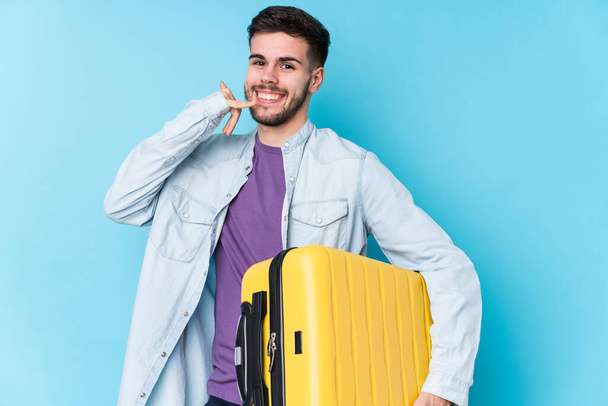 młody biały podróżnik człowiek trzymając walizkę odizolowany pokazując telefon komórkowy gest z palcami. - Zdjęcie, obraz