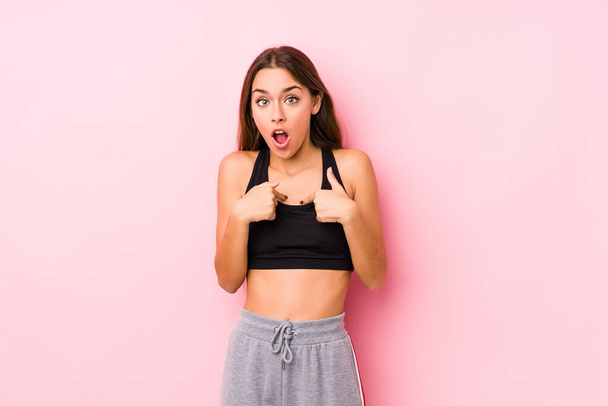 Joven mujer de fitness caucásica posando en un fondo rosa sorprendido señalando con el dedo, sonriendo ampliamente. - Foto, Imagen