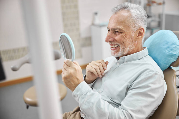 Gladsome aged man looking at his teeth in the mirror - Photo, image