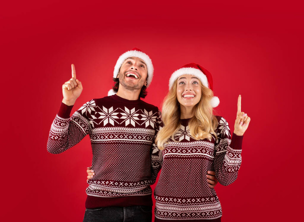 Vánoční prodej. Šťastný milennial pár v Santa klobouky ukazuje nahoru na červeném pozadí - Fotografie, Obrázek