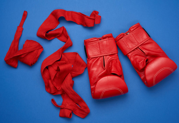 пара красных кожаных боксерских перчаток и текстильно-красная эластичная повязка для рук лежат на синем фоне, спортивный комплект, плоский уголок - Фото, изображение