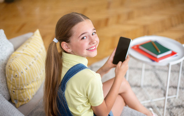 Dívka pomocí mobilu sedí na gauči a ohlíží se - Fotografie, Obrázek