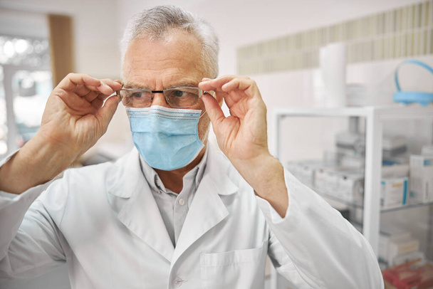 Médico profesional en una máscara quirúrgica tocando sus gafas - Foto, imagen