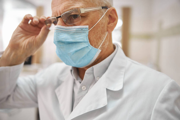 Старший мужчина в маске и очках работает в клинике - Фото, изображение