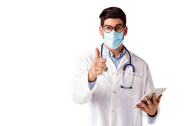 Shot of male doctor giving thumbs up and wearing face mask while standing at isolated white background.  - Foto, Imagen