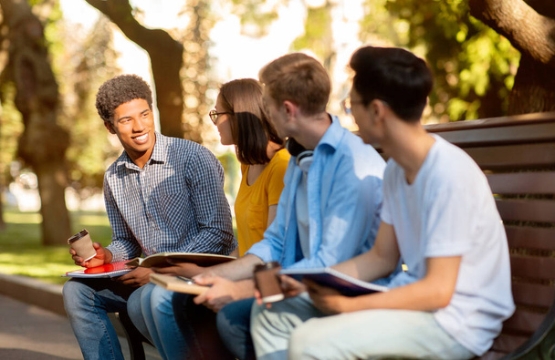 Vier Gymnasiasten diskutieren auf Bank im Park über Unterricht - Foto, Bild