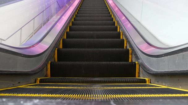 低角度の近代的なエスカレーターの階段の視点をループ化した。自動エレベーター機構。紫色の光で照らされた階段の上の黄色の線。未来的な空の機械階段がまっすぐ動く. - 写真・画像