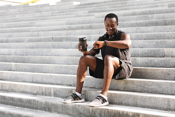 Afrikkalainen Athete Guy tarkistaminen Fitness Tracker harjoittelun jälkeen ulkona, istuu portailla - Valokuva, kuva
