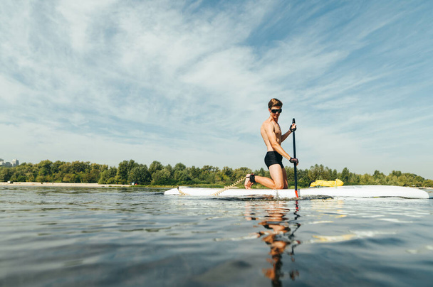 Handsome young man with a sports torso calmly paddles on a sup board on the river. The guy swims on the water on a paddle board. Copy space - Fotoğraf, Görsel