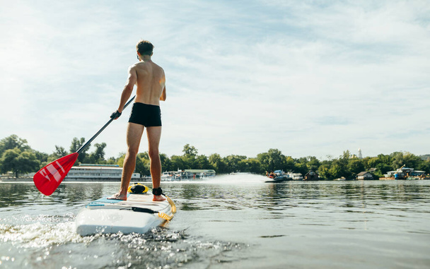 Atletický mladý muž plave podél řeky na pádlovém prkně, pohled zezadu. Sportovní vlaky na sup palubě, aktivně pádluje - Fotografie, Obrázek