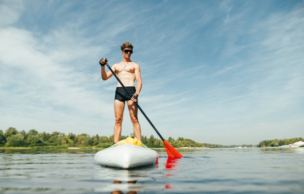 Pohledný mladý muž stojící na prkně s pádlem v ruce na vodě. Aktivní odpočinek na veslařské palubě na řece. Chlápek plave po řece na sup boardu - Fotografie, Obrázek