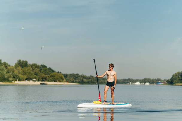 Athletic guy with a beautiful muscular summer stands on the water on a paddle board with an oar in his hand and looks away at the copy space. Swimming on a sup board - Photo, Image