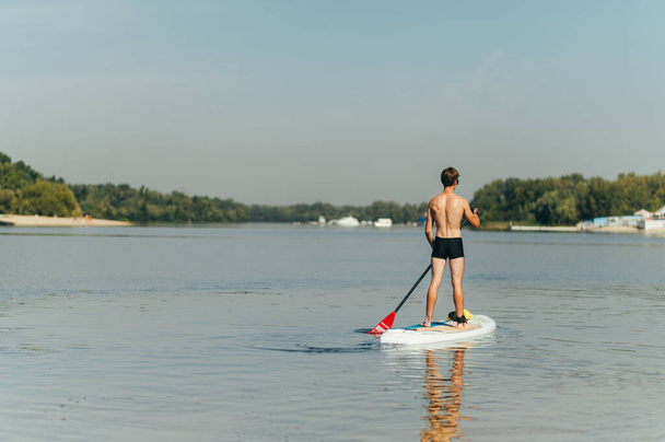 Atletik genç adam nehir kıyısındaki bir panoda kürek çekiyor, arkadan görüyor. Sıcak bir günde kürek tahtasında antrenman yapmak. - Fotoğraf, Görsel