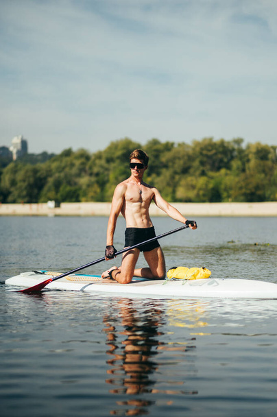 筋肉運動の若い男は夏に川で自由な時間を過ごします,アップボード上のパドル.晴れた日にボード上のワークアウト。垂直 - 写真・画像