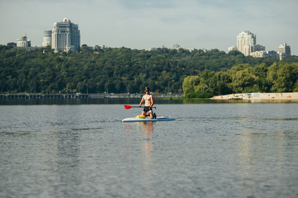 Muskularny sportowiec spędza wolny czas latem nad rzeką, wiosła na desce. Trening na desce w słoneczny dzień. - Zdjęcie, obraz