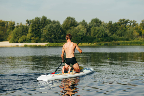 Задняя часть молодого человека плавает на доске на озере на фоне пляжа и деревьев, активно гребя. Активный отдых на воде. - Фото, изображение