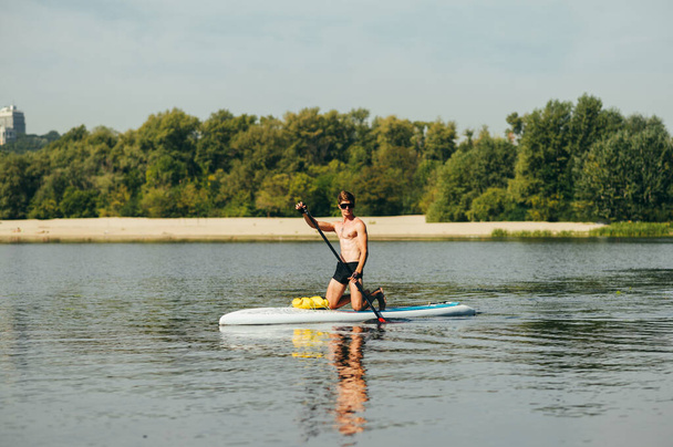Handsome young man rides on the river on a sup board, paddles and looks at the camera. Athlete trains on a sup board on a sunny summer day - Photo, Image