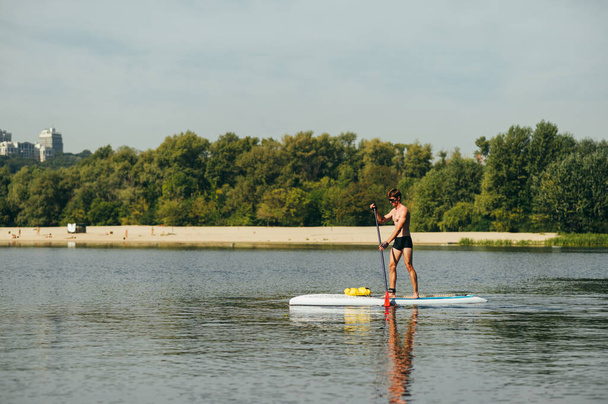 Atletik genç adam nehirde sup tahtasının üzerinde duruyor ve bir kumsal ve ağaçların arka planında kürek çekiyor. Nehirde dinlen.. - Fotoğraf, Görsel