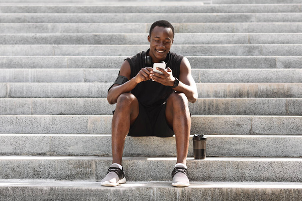 Тренировка. Спортсмен черный парень отдыхает на ступеньках со смартфоном - Фото, изображение