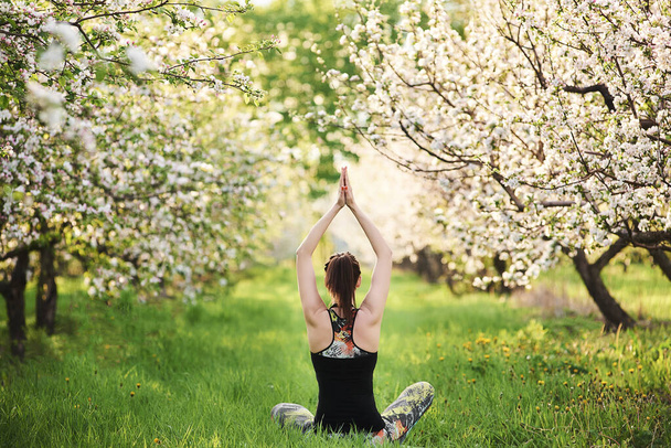 Yogi fille assise sur la pelouse verte levant les mains vers le haut effectuer Namaste, vue arrière, pleine longueur, vue arrière. Concept de vie saine et équilibre naturel entre le corps et la nature. - Photo, image