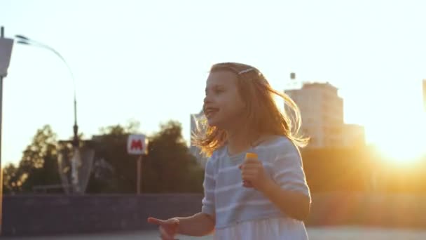 1人のアクティブな小さな就学前の女の子は、通りで余暇を過ごし、喜びでジャンプし、離れて見て、指で横に方向を示し、カメラを見て吹いているシャボン玉を保持し、子供時代の概念 - 映像、動画