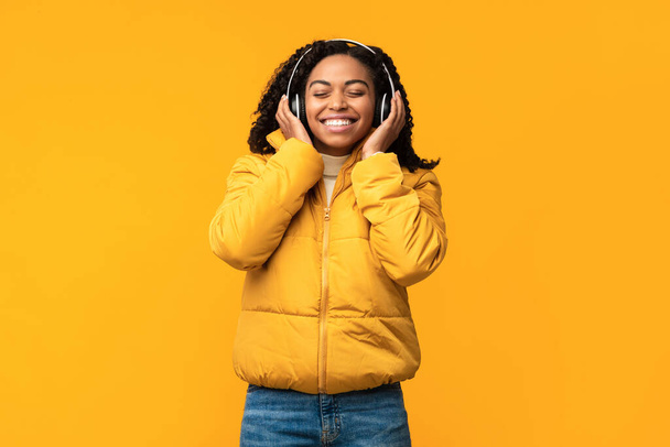 Zwarte vrouw luisteren muziek over gele achtergrond, dragen van winterkleding - Foto, afbeelding