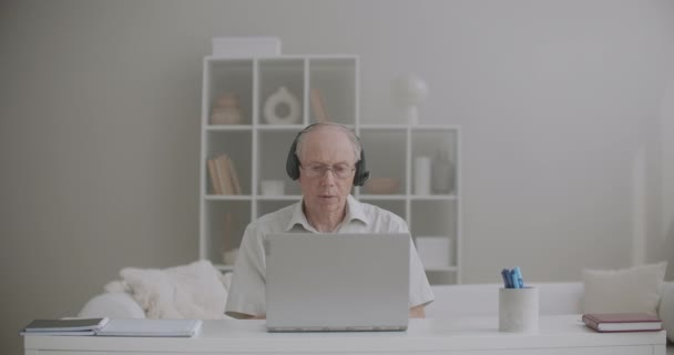 高齢の男性教師は講義をオンラインで行い家にいますインターネット接続とヘッドフォンでノートパソコンを使います - 映像、動画