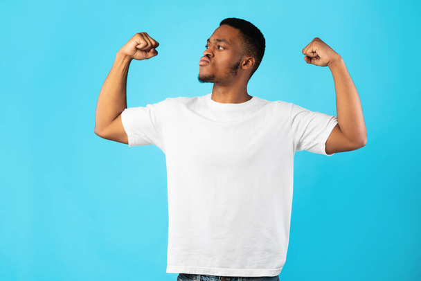 Silný africký americký muž ukazuje biceps stojící nad modrým pozadím - Fotografie, Obrázek