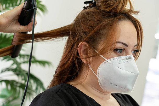Elegant young hairdresser with face mask straightening the hair of a red hair client's in a modern hairdressing salon . Social distancing. New normality - Photo, Image