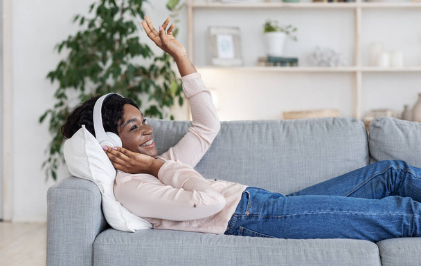 Radosna afrykańska dama ciesząca się muzyką w słuchawkach bezprzewodowych podczas relaksu na kanapie - Zdjęcie, obraz