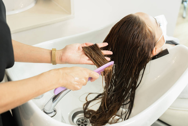 Fiatal lány hosszú barna hajjal, fésülködik és bogozza a haját a fodrásznál. Társadalmi távolság. Használd az arcmaszkot. Közelkép - Fotó, kép