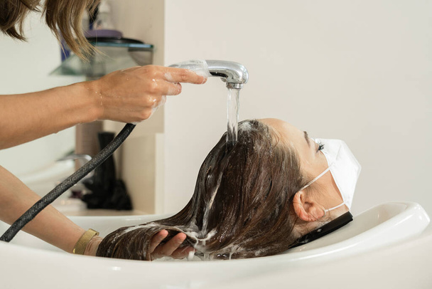 Young girl with long brown hair, rinsing her head in the hairdresser's. Social distancing. Use of the face mask. Close-up - Photo, Image
