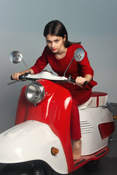  Κορίτσι με κόκκινο κοστούμι. Το κορίτσι κάθεται πάνω σε ένα κόκκινο μοτοποδήλατο. Φωτογραφία μόδας. - Φωτογραφία, εικόνα