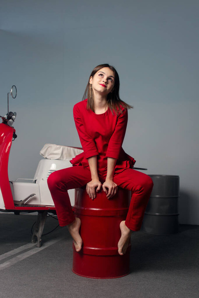  young woman in a red suit. The young woman is sitting on a red barrel. In the background, a red moped. - Valokuva, kuva