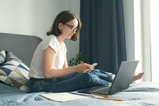 Young business woman, teacher, student working or studying online on her bed with mobile phone and black laptop. Distance work from home or online education concept. Selective focus - Photo, image