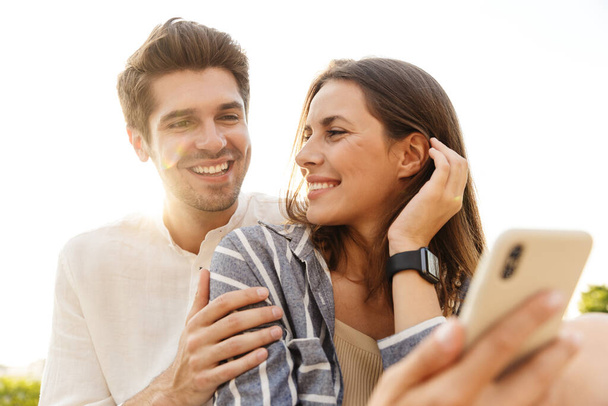 Image of young caucasian happy couple smiling and hugging while sitting outdoors with cellphone - Φωτογραφία, εικόνα