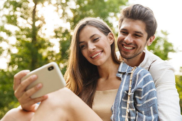 Image of young caucasian happy couple smiling and hugging while holding cellphone in park - Φωτογραφία, εικόνα