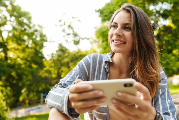 Image of young caucasian brunette woman smiling and holding cellphone while walking in park - Φωτογραφία, εικόνα